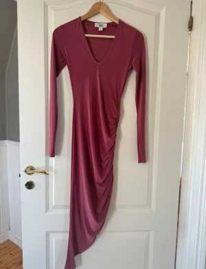 Rebecka Stella. Lång klänning med drapering - Säljer en elegant vinröd långklänning från Rebeccas Stellas äldre kollektion med v-ringad hals och långa ärmar. Klänningen har snygg drapering på sidan som ger en cool asymmetrisk look. Materialet är mjukt och stretchigt.