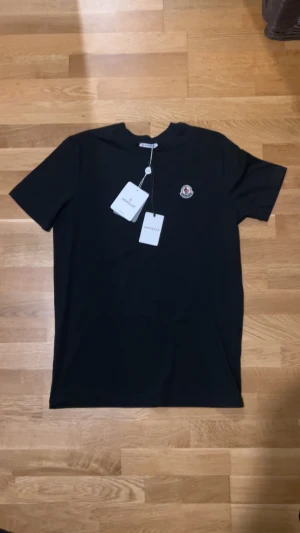 Svart Moncler t-shirt med logga - Svart t-shirt från Moncler i klassisk modell med rund hals och korta ärmar. På bröstet sitter Monclers ikoniska broderade logga i vitt, rött och blått. T-shirten är gjord i mjuk bomull och har en clean, stilren look som funkar till allt.