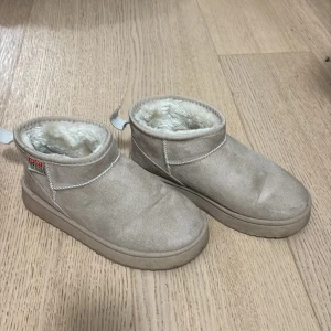 Beige boots med pälsfoder - Mysiga beige boots med rund tå och platt sula, fodrade med mjuk vit päls på insidan. Utsidan är i mocka och har en enkel design med synliga sömmar och en praktisk flik bak för att lätt dra på dem. 