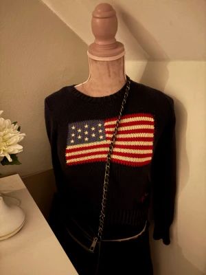 Mörkblå stickad tröja Polo Ralph Lauren (ordinarie pris 5000) - Snygg mörkblå stickad tröja från Polo Ralph Lauren med amerikansk flagga i rött, vitt och blått på bröstet. RL-broderi i rött vid nederkanten. Rund hals och långa ärmar, perfekt för dig som gillar klassisk college-stil.