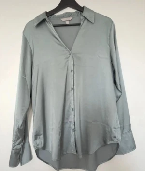  satinliknande skjorta H&M - Snygg skjorta från H&M i ett glansigt, satinliknande material. Skjortan har klassisk krage, knappar framtill och långa ärmar. Ge färg till hösten med denna underbart fina färg!   Använd 2 gånger. Har en likadan i annan färg som är en favorit väldig skön!  FÄRGEN ÄR TURKOS/BLÅGRÅ Djur och rökfritt hem 🏠