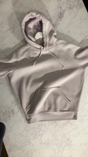 Ljuslila hoodie med magficka - En stilren ljuslila hoodie med klassisk magficka och dragsko i huvan. Hoodien har raglanärmar och ribbade muddar vid ärmslut och nederkant. Perfekt för en chill och avslappnad look. Materialet känns mjukt och skönt mot huden.