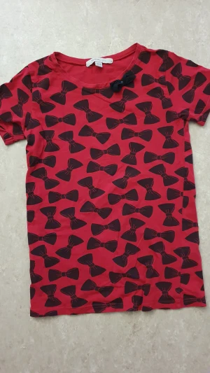 T-shirt - Röd t-shirt från Mywear Young i storlek 146/152. T-shirten har korta ärmar, svart rosettprint över hela och en extra svart tygrosett vid halsen. Tillverkad i mjuk bomull, perfekt för dig som gillar lekfull stil.