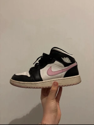 Nike Air Jordan 1 Mid svart/vit/rosa - Säljer ett par Nike Air Jordan 1 Mid med svart och vitt skinn, rosa swoosh och matchande rosa detalj på plösen. Klassisk rund tå, snörning och platt sula. Perfekta för dig som vill sticka ut med en unik färgkombo och ikonisk siluett.