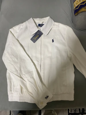 Vit overshirt från Polo Ralph Lauren - Snygg vit overshirt från Polo Ralph Lauren med klassisk krage, broderad logga på bröstet och elastisk nederkant. Jackan har dragkedja framtill och knappar vid ärmslut med Polo-broderi. Perfekt för dig som gillar stilrena och tidlösa plagg.