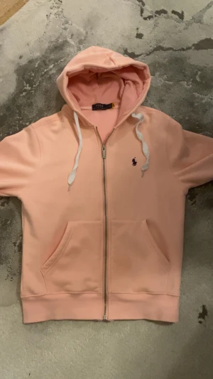 Ljusrosa hoodie från Polo Ralph Lauren - Snygg ljusrosa hoodie från Polo Ralph Lauren med dragkedja och huva med vita snören. Tröjan har två fickor framtill och ett broderat mörklila Polo-logo på bröstet. Insidan är mjuk och fleecefodrad för extra komfort. Perfekt för chill dagar.