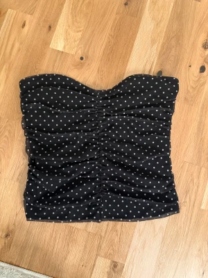 Svart prickig tubtopp - Säljer en svart tubtopp med vita prickar och snygg rynkdetalj framtill. Toppen är axelbandslös och har en bandeauformad överdel som sitter tight. Perfekt att styla med jeans eller kjol för en cool look. Storlek M men passar mindre också 