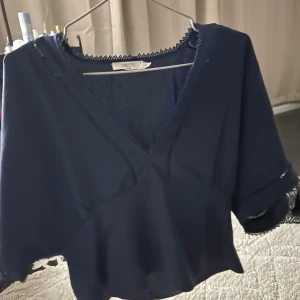 Mörkblå blus med v-ringning från Milos - Säljer en mörkblå blus från Milos med v-ringning och dekorativa spetsdetaljer längs halsringningen och ärmsluten. Blusen har korta, vida ärmar och en croppad passform som ger en trendig siluett. Perfekt för dig som gillar stilrena och snygga toppar.