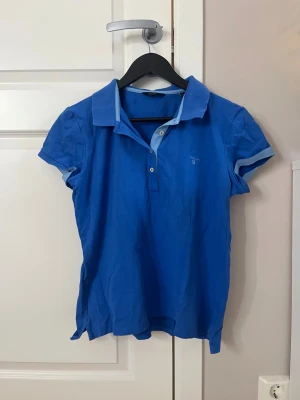 Blå pikétröja från GANT, storlek L - Snygg blå pikétröja från GANT i storlek L. Tröjan har klassisk krage och knappslå med två knappar, samt ljusblå detaljer på krage och ärmslut.