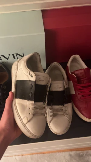 ÄKTA Valentino Garavani sneakers - Säljer ett par vita Valentino Garavani sneakers i skinn med bred svart rem över mitten. Klassisk rund tå, platt sula och snörning framtill. Märkeslogga på plösen. Snygga och stilrena skor som lyfter din outfit. Storlek 40 men passar lite större. Endast skona medföljs