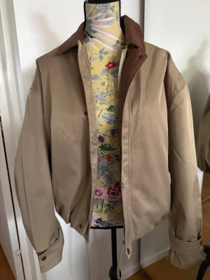 Beige bomberjacka Acne Studios - Snygg beige bomberjacka från Acne Studios med brunt skinnkrage och detaljer. Jackan har dragkedja och tryckknappar framtill, samt en patch med Acne Studios Stockholm 1996 på ryggen. Oversized passform och långa ärmar, perfekt för en clean och modern look.