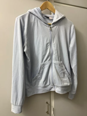 Ljusblå velour hoodie från Gina Tricot - Säljer en ljusblå velour hoodie från Gina Tricot i mjuk polyester. Tröjan har huva, dragkedja framtill och två stora fickor. Perfekt för chill dagar och enkel att matcha med jeans eller mjukisbyxor. Långärmad och med normal passform. Snören saknas till luvan.