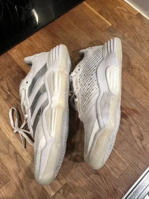 Adidas stabil handbollsskor, vita med silverdetaljer - Säljer ett par vita Adidas stabil handbollsskor med silvriga ränder på sidorna. Skorna har en chunky sula och är tillverkade i mesh och syntetmaterial 