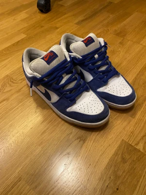 Nike SB Dunk Low Pro Deep Royal Blue - Nike SB Dunk Low Pro PRM i färgen Deep Royal Blue. Skorna har vit mesh och blå mocka med blå snörning, röd Nike-logga på tungan och hälen samt perforerad tå. Låg modell med platt sula och klassisk Dunk-silhuett. Perfekt för dig som gillar streetwear och skate.