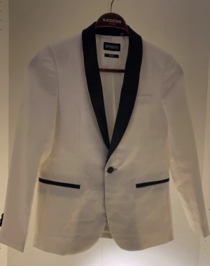 Vit kavaj med svarta detaljer Opposuits - Snygg vit kavaj från Opposuits med svarta slag och svarta detaljer vid fickorna. Klassisk passform med en knapp framtill och långa ärmar. Perfekt för dig som vill sticka ut med en stilren och modern look.