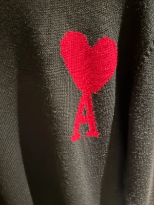 Svart stickad tröja från Ami - Svart stickad tröja från Ami med röd hjärtapplikation och bokstaven A på bröstet. Tröjan har rund hals med ribbad kant och långa ärmar. Perfekt för dig som gillar stilrena plagg med en twist. Materialet är mjukt och håller värmen.