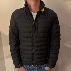 Svart dunjacka Parajumpers herr - Svart dunjacka från Parajumpers med klassisk gul detalj vid kragen och patch på ärmen. Jackan har quiltad design, dragkedja framtill och hög krage. Perfekt för kalla dagar och har en snygg, normal passform. Materialet är polyester och dun.