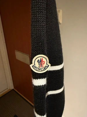 Svart stickad Moncler tröja med ränder - Svart stickad tröja från Moncler med vita ränder på ena ärmen och klassisk Moncler-logga. Tröjan har rund hals, ribbade muddar och detaljerad stickning vid axlarna. Perfekt för dig som gillar stilrena märkesplagg med snygga detaljer.