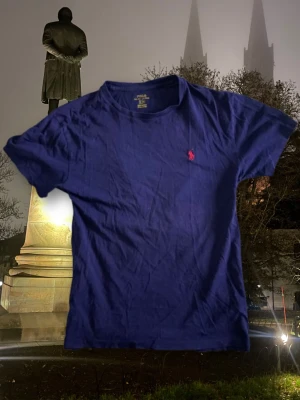 Blå t-shirt från Polo Ralph Lauren - Snygg blå t-shirt från Polo Ralph Lauren med klassisk röd logobroderi på bröstet. Modellen är kortärmad och har en rund hals. Tillverkad i mjuk bomull som känns skön mot huden. Perfekt för dig som gillar stilrena och ikoniska plagg.