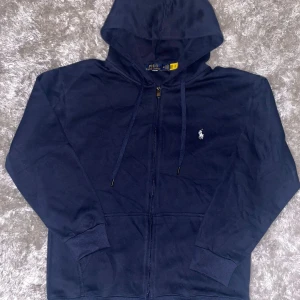 Marinblå hoodie från Polo Ralph Lauren - Snygg marinblå hoodie från Polo Ralph Lauren med dragkedja och huva. Klassisk vit broderad logga på bröstet, ribbade muddar och fickor framtill. Perfekt för dig som gillar stilren och sportig look. Helt ny