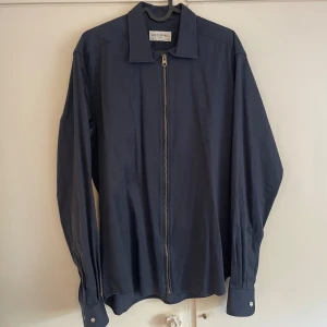 Marinblå overshirt från John Henric - Snygg marinblå overshirt från John Henric med dragkedja framtill och klassisk krage. Skjortan har långa ärmar med manschettknappar och är tillverkad i ett skönt bomullstyg. Perfekt för dig som vill ha en stilren och modern look. Nypris 899kr.