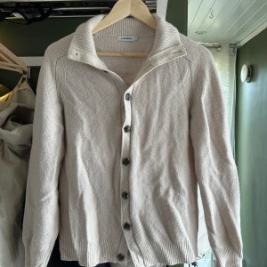 kofta från J.Lindeberg - Snygg beige stickad kofta från J.Lindeberg med klassisk krage och knappar hela vägen framtill. Koftan har långärmade ribbade muddar och är tillverkad i mjukt stickat material som ger en chill och stilren vibe. Perfekt för lager-på-lager under hösten och vintern. Det är en L men den sitter som en S, så har du storlek S i tröjor är den perfekt!