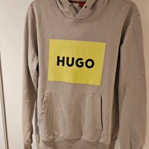Ljusgrå hoodie från HUGO med tryck - Snygg ljusgrå hoodie från HUGO med stor gul logga framtill. Tröjan har huva, känguruficka och är tillverkad i mjuk bomull. Perfekt för en chill och stilren look. Långärmad och loose fit för maximal komfort.