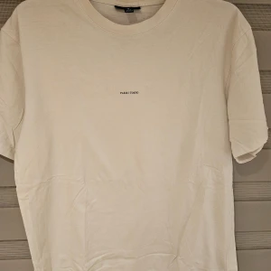 Vit t-shirt från Icono Couture M - Snygg vit t-shirt från Icono Couture med tryck 'PARIS / TOKYO' på bröstet och stor logga på ryggen. Modellen är relaxed fit med klassisk rund hals och korta ärmar. Perfekt för en clean och stilren look.