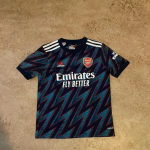 Arsenaltröja med White på ryggen - Arsenal bortatröja från Adidas med spelarnamn White och nummer 4. Tröjan är marinblå med blixtmönster i blått och röda detaljer, klassiska vita Adidas-ränder på axlarna och broderat Arsenal-märke på bröstet. Storlek: 142-150. Jag har tagit bra hand om tröjan men det syns att den är använd. Säljer då den har blivit för liten. Köpte för runt 1000