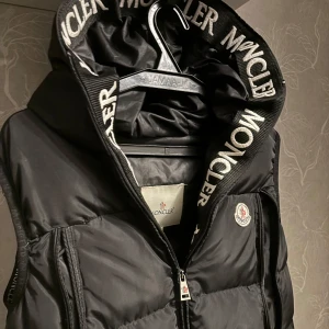 Svart dunväst från Moncler med huva - Svart dunväst från Moncler med stor huva och vit logotyptext längs kanten. Västen har två dragkedjeförsedda fickor, quiltad design och Moncler-märke på bröstet. Snygg och sportig look, perfekt för kyliga dagar. Dragkedja framtill och stilren passform. I väldigt bra skick. Strl M (damstorlek skulle ja säga)