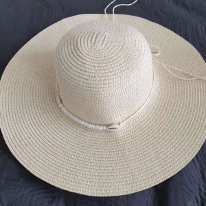 Beige solhatt med pärldetalj - Snygg beige solhatt med breda brätten och dekorativt band med pärlor och snäckor runt kronan. Hatten är flätad och har justerbart band på insidan för perfekt passform. 