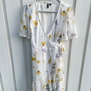 Vit blommig omlottklänning Vero Moda - Superfin vit omlottklänning från Vero Moda med blommigt mönster i gult och lila. Klänningen har korta ärmar, volangdetaljer och knyt i midjan. Perfekt för varma dagar och har en luftig känsla. Säljes i storlek M.