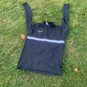 Svart Nike Dri-Fit träningströja M - Svart långärmad träningströja från Nike med vit logga och en reflekterande horisontell rand över bröstet. Tröjan har half zip och är tillverkad i lätt och snabbtorkande polyester. Perfekt för löpning eller gymmet.
