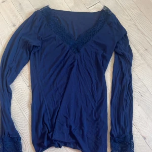 Mörkblå långärmad intimissimi spetsblus S - Mörkblå långärmad blus med v-ringning och fina spetsdetaljer både vid halsringningen och ärmsluten. Toppens material är mjukt och lätt, perfekt för en stilren look. Säljs i storlek S.