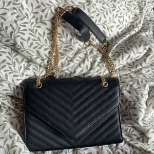Svart quiltad axelväska med guldkedja - Snygg svart axelväska i quiltad design med guldfärgade kedjor och detaljer. Väskan har ett klassiskt kuvertformat lock och insida med dragkedjefack. 