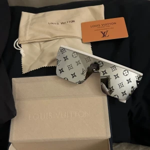 vita monogram solglasögon  - Snygga solglasögon med vita bågar och spegelglas med ikoniskt monogrammönster. Sköldpaddsinspirerad shield-form och breda skalmar med logotyp. Perfekt statement piece för dig som vill sticka ut.