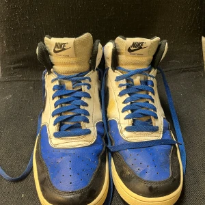 Nike Court Vision Mid blå/vita sneakers - Nike Court Vision Mid sneakers i blått, vitt och svart skinn med klassisk svart swoosh. Högt skaft, blå snörning och beige sula ger en retro vibe. Perforerad tåbox för extra ventilation och cool look. Perfekta för dig som gillar streetstyle och sportig känsla. Allt är bra förutom att ena skon har en gräsfläck. 