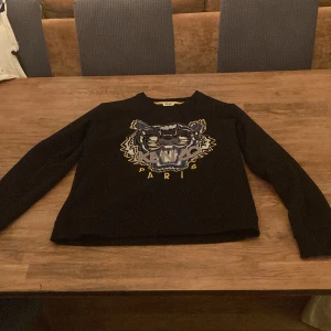 Svart Kenzo tröja med tigerbrodyr - Svart långärmad tröja från Kenzo med ikonisk broderad tiger och texten 'Kenzo Paris' i gult och blått framtill. Rund halsringning med gul detalj på insidan. Perfekt statement piece för dig som gillar streetstyle.