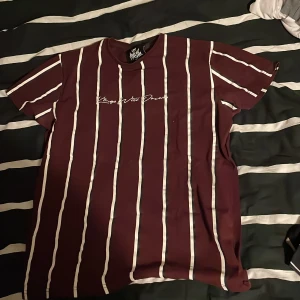 Randig vinröd t-shirt Kings Will Dream - Snygg vinröd t-shirt från Kings Will Dream med vita vertikala ränder och broderad logga på bröstet. Klassisk rund hals och korta ärmar. Perfekt för dig som gillar streetwear och vill sticka ut med en fräsch design.