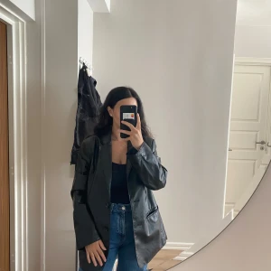 Svart skinnkavaj från H&M - Snygg svart kavaj i skinnimitation från H&M, storlek S. Jackan har klassisk kavajkrage, två framfickor och knäppning framtill. Passformen är loose och den har långa ärmar. Perfekt för dig som gillar en cool och trendig look.