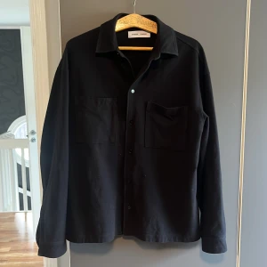 Svart overshirt från Samsøe Samsøe - Stilren svart overshirt från Samsøe Samsøe med klassisk krage, två stora bröstfickor och knäppning framtill. Skjortan har lång ärm och en rak, lite loose passform. Perfekt att slänga över en t-shirt för en clean look. Storlek S/M