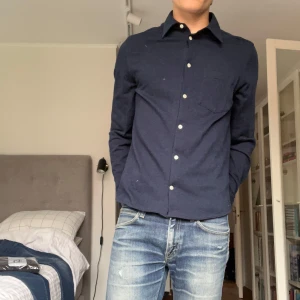 Mörkblå skjorta från J.Lindeberg - Stilren mörkblå skjorta från J.Lindeberg.     100% bomull. klassisk krage, vita knappar och bröstficka. Skjortan är långärmad och har en normal passform