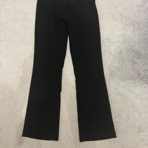 Svarta bootcut kostymbyxor - Snygga svarta kostymbyxor med bootcut-snitt och låg midja. Byxorna har klassiska hällor för bälte och stängs med knapp och dragkedja. Perfekta för dig som gillar en stilren look med lite utsvängda ben. Materialet känns mjukt och har en lätt stretch.