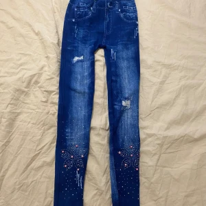 Blå skinny jeans med pärldetaljer - Snygga blå skinny jeans med slitningar och coola pärldetaljer i rosa och silver nertill på benen. Jeansen har dekorativa sömmar, hjärtformade bakfickor och ett blommigt mönster av pärlor framtill. Perfekta för dig som gillar att sticka ut lite extra.