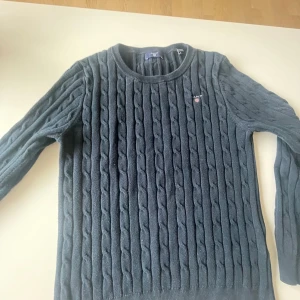 Mörkblå kabelstickad tröja från Gant - Säljer en klassisk mörkblå kabelstickad tröja från Gant med rund halsringning och broderad logga på bröstet. Tröjan är långärmad och har en tidlös design som passar perfekt till jeans eller kjol.