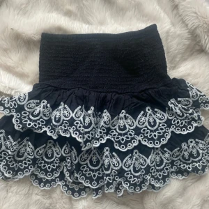 Svart volangkjol med broderad spets - Supersöt svart kortkjol med bred smock i midjan och två lager volanger med vit broderad spetskant. Kjolen har en feminin och lekfull vibe, perfekt till sommaren. Materialet känns lätt och luftigt, och spetsdetaljerna gör den extra unik.