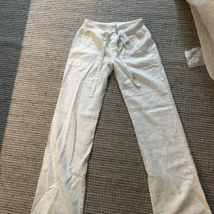 Vita linnebyxor från Vero Moda - Snygga lågmidjade! vita linnebyxor från Vero Moda med bred resårmidja och snörning framtill. Byxorna har raka ben/ vida, två fickor fram och dekorativa fickor bak. 