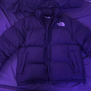 Svart dunjacka The North Face 700 - Säljer en svart dunjacka från The North Face, modell 700. Jackan är riktigt puffig med hög krage, dragkedja framtill och två stora fickor. Klassisk logga på bröstet och ärmen. Perfekt för kalla dagar när du vill hålla värmen och ändå se clean ut.