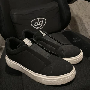 Svarta slip-on sneakers med vit sula - Stilrena svarta slip-on sneakers med tjock vit sula och minimalistisk design. Ovandelen är i mjukt tygmaterial och skon har en rund tå. Perfekta för dig som gillar enkel och clean stil utan snörning.