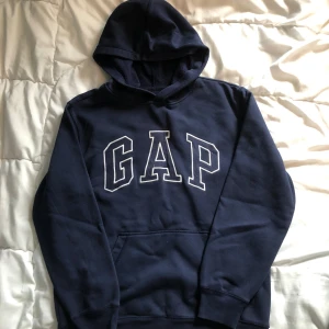 Mörkblå GAP hoodie  - Mörkblå hoodie från GAP med stor vit logga framtill. Klassisk modell med huva och känguruficka. Mjuk insida och ribbade muddar vid ärmslut och nederkant. Perfekt för chill dagar eller när du vill ha en enkel och snygg look.
